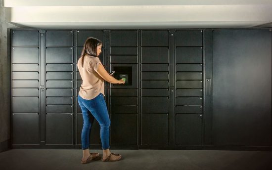 Mulher usando camisa clara e calça jeans, estende os braços para abrir a porta do armário inteligente do condomínio. O armário tem cor grafite escuro.